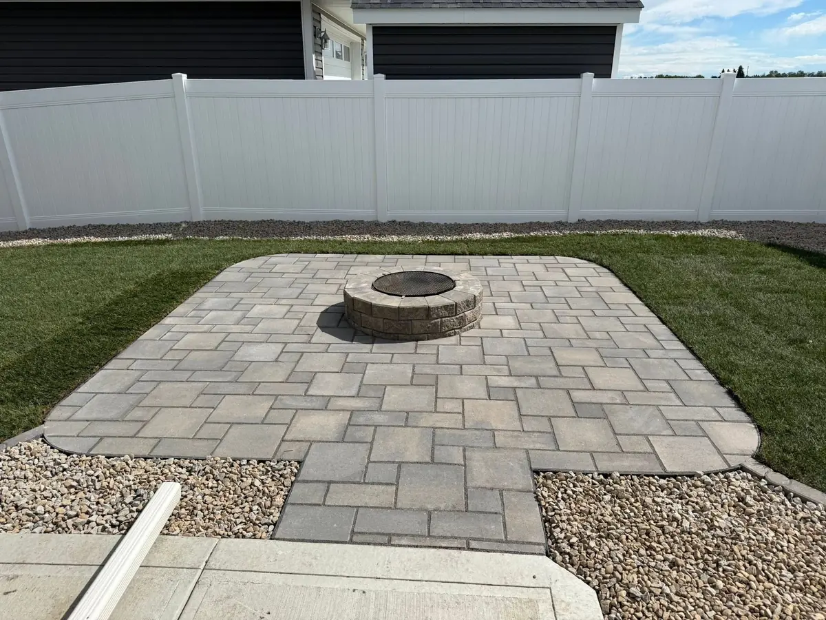 A paver patio with a stone fire pit, set into fresh sod behind a white vinyl fence